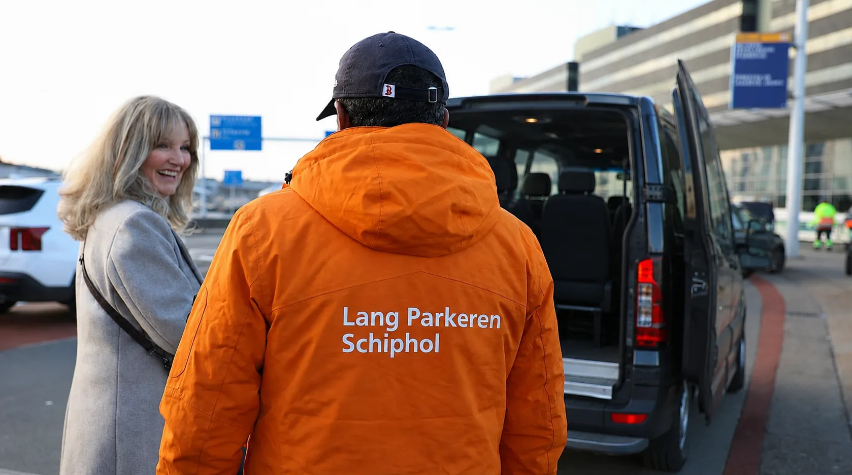 Chauffeur van Lang Parkeren Schiphol in oranje bedrijfsjas begeleidt een reizigster naar de shuttlebus bij de vertrekhal op Schiphol.
