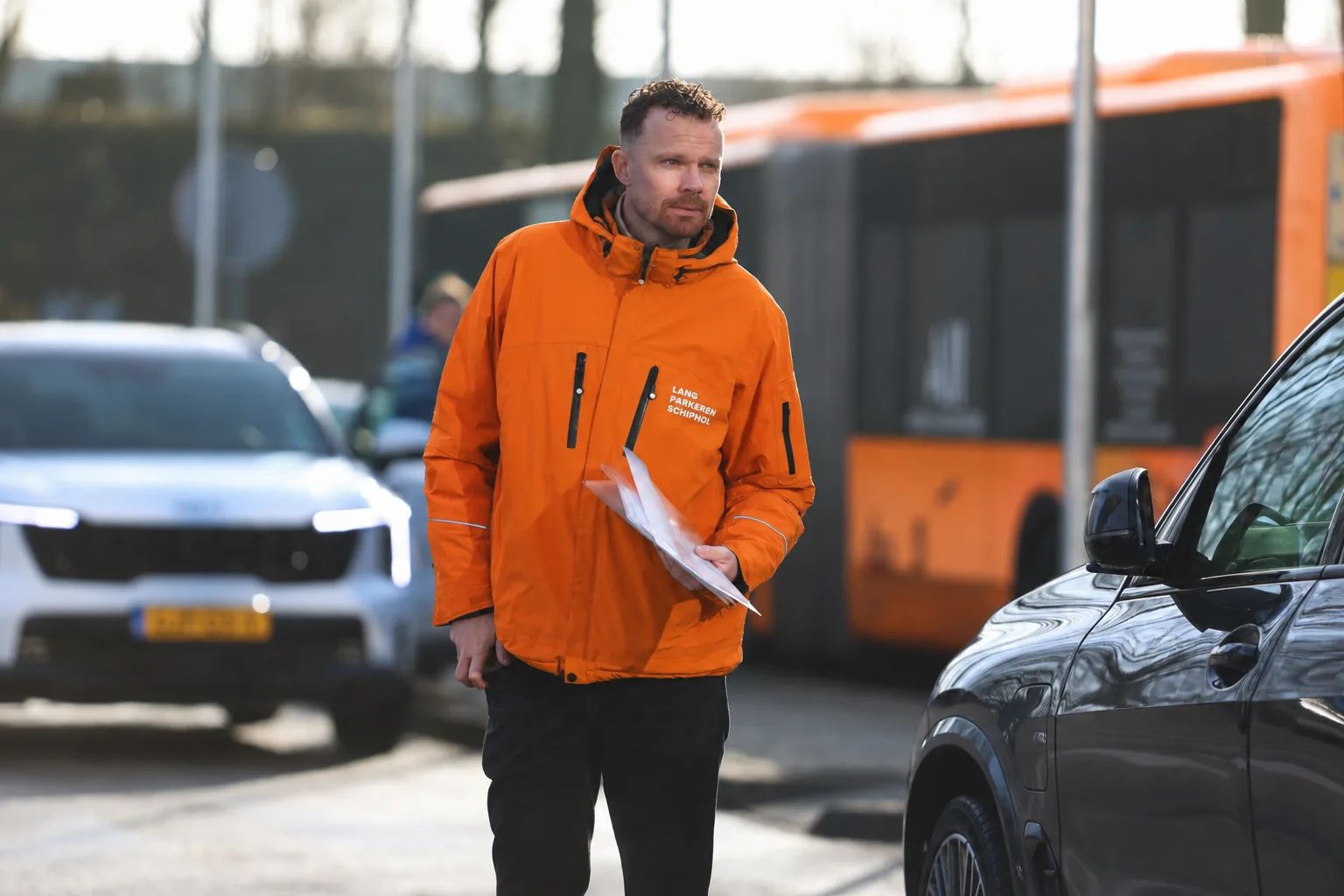 Chauffeur van Lang Parkeren Schiphol in oranje bedrijfsjas met de papieren van een reservering in zijn hand, op het parkeerterrein met de shuttlebus op de achtergrond.