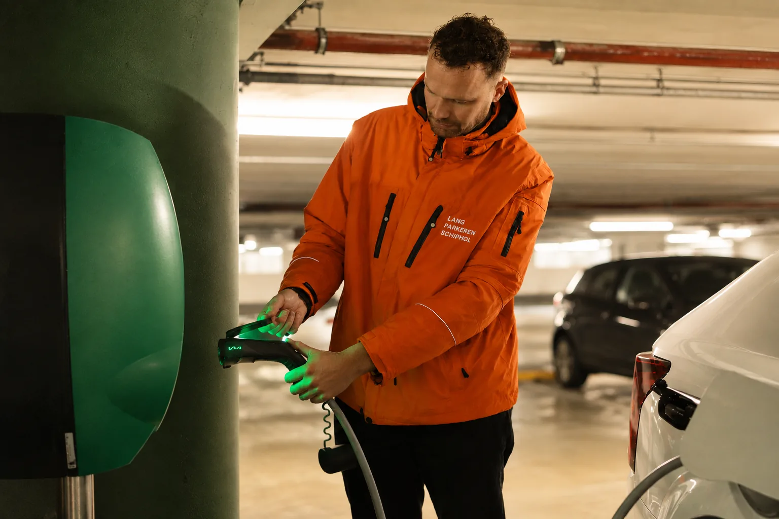 Medewerker van Lang Parkeren Schiphol sluit een laadkabel aan op een elektrische auto bij een laadpunt in de overdekte parkeergarage.
